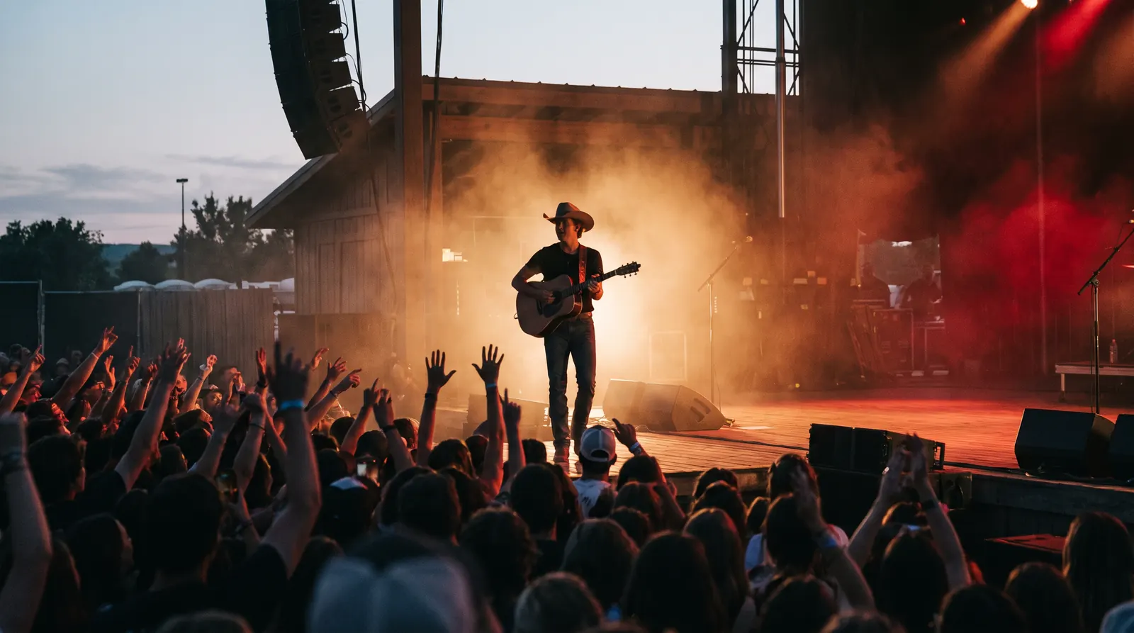 Country singer-guitarist silhouetted against amber stage lights at an outdoor amphitheater, hands raised in the crowd below